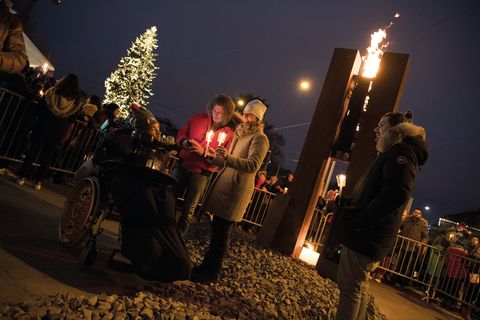 Eine Skulptur mit brennender Flamme bei Nacht, davor Menschen mit Kerzen und ein Weihnachtsbaum